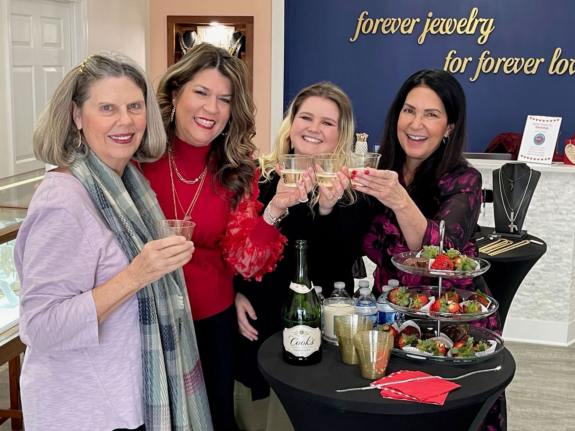 Four women toasting with champagne near a table with strawberries and jewelry in a shop.