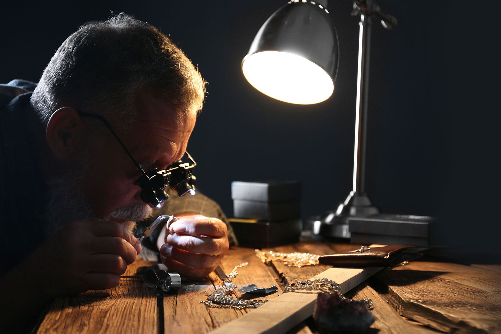 Jeweler using magnifying glasses under a desk lamp, examining small objects.