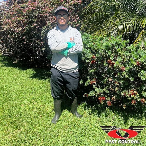 A man is standing in the grass with his arms crossed in front of a bush.