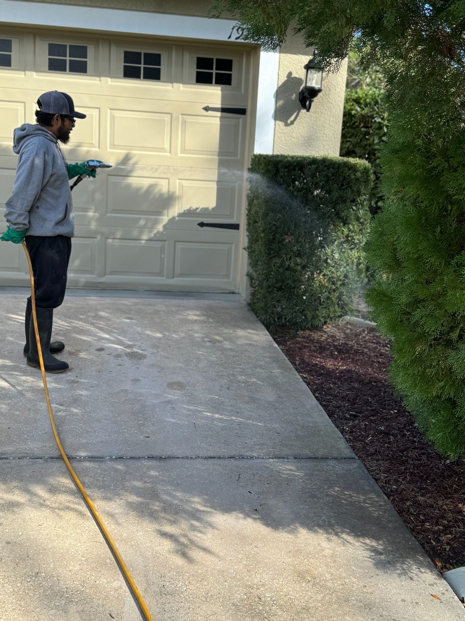 A man is spraying a hose on a driveway in front of a garage door.