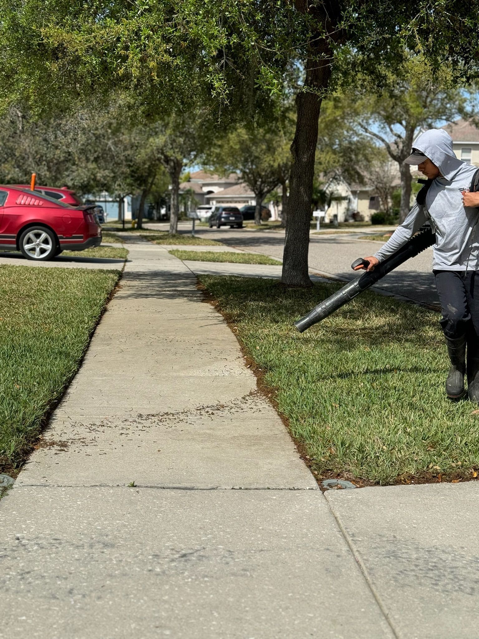 A man is blowing leaves on the sidewalk next to a red car.