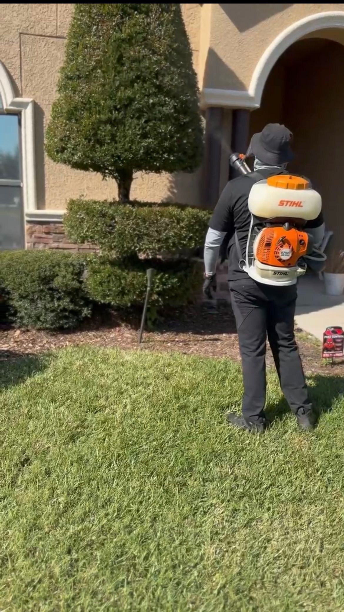 Person spraying lawn with a backpack sprayer in front of a house.