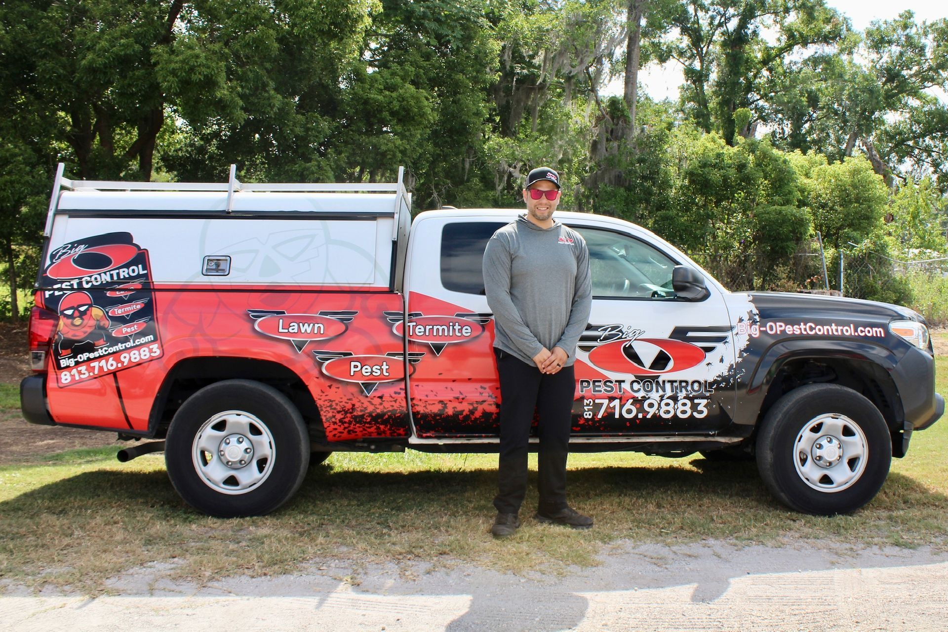 Man standing beside his company truck. Truck is black, white, and red. Trees in the background.