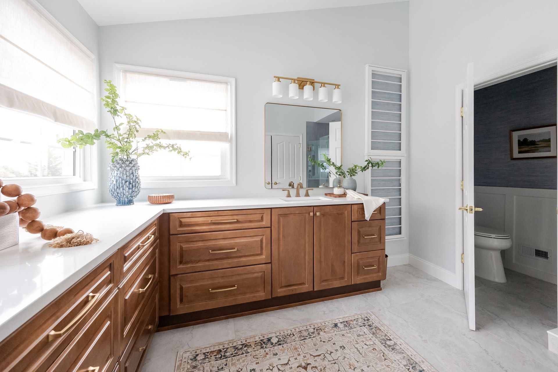 A bright bathroom with a wood vanity, white countertops, two windows, and an open door leading to a separate toilet area.