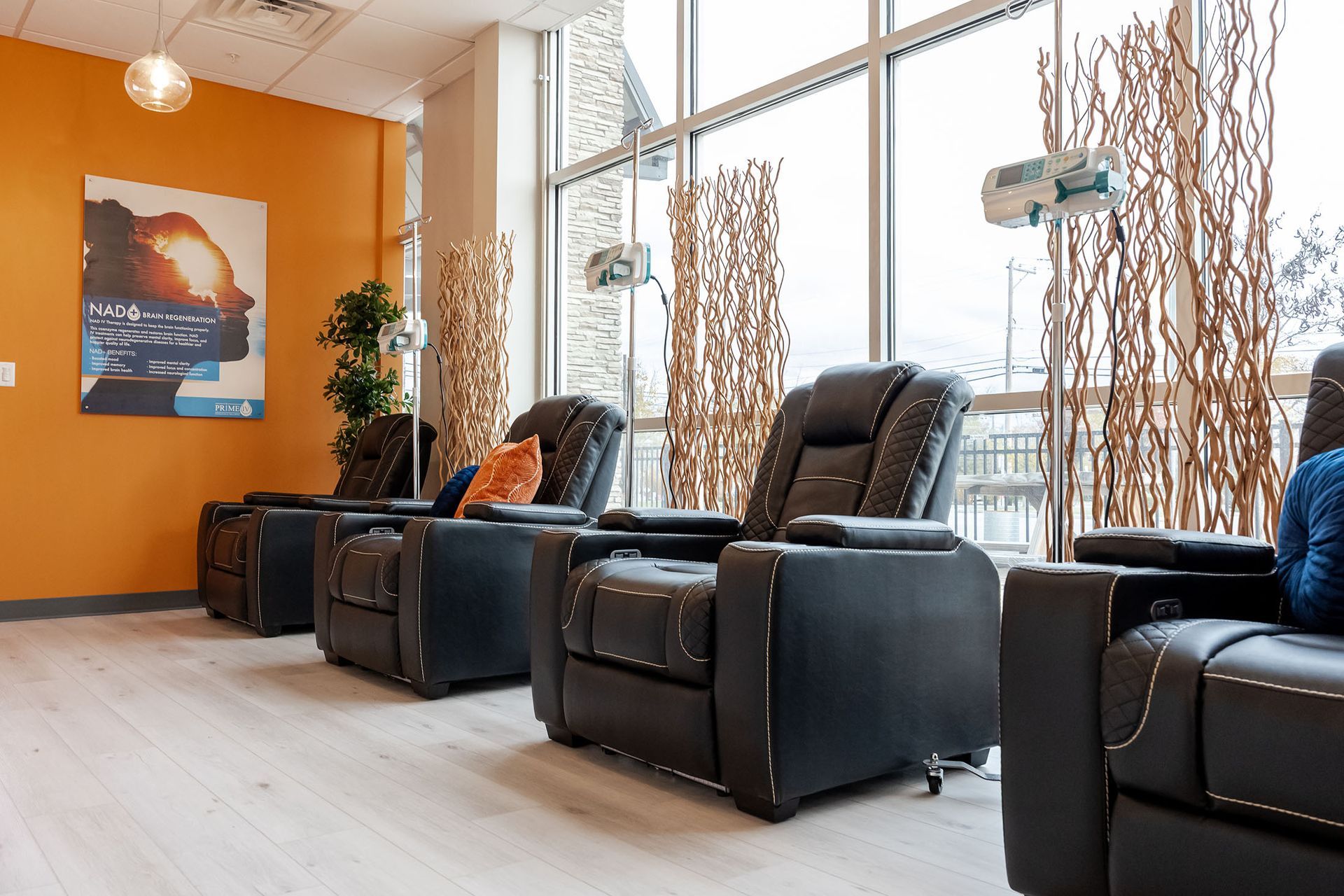 Modern waiting room with black recliners, orange accent wall, tall windows, and decorative branches.