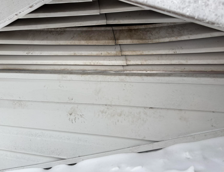 Gable vent with dark staining and sagging horizontal slats above a wall section covered by snow.