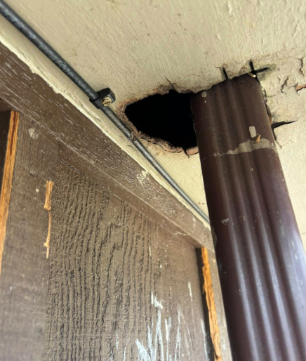 A dark hole in a beige ceiling beside a brown metal downspout, with an exposed electrical wire running nearby.