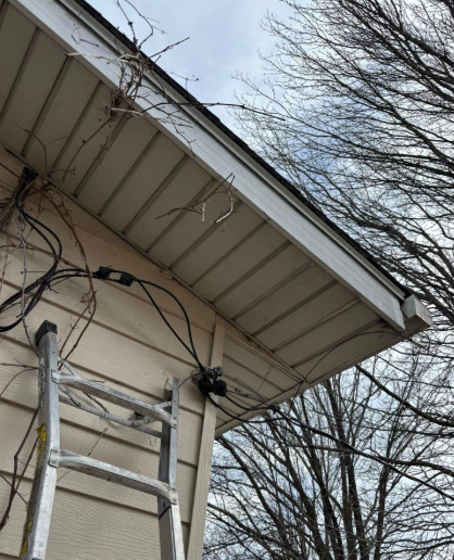 A metal ladder leans against the exterior of a house, where dark cables are attached near the roofline and soffit.