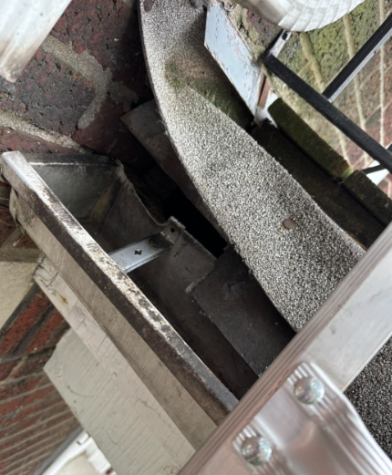 A close-up view from a ladder showing an empty, dirty residential gutter and a shingled roof edge against a brick wall.