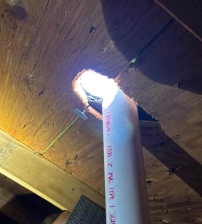 A white PVC plumbing vent pipe extending through a hole in plywood attic sheathing, showing daylight from outside.