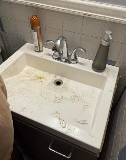 A dirty white bathroom sink with chrome faucets, a brown spray bottle, and a dark grey soap dispenser on the counter.