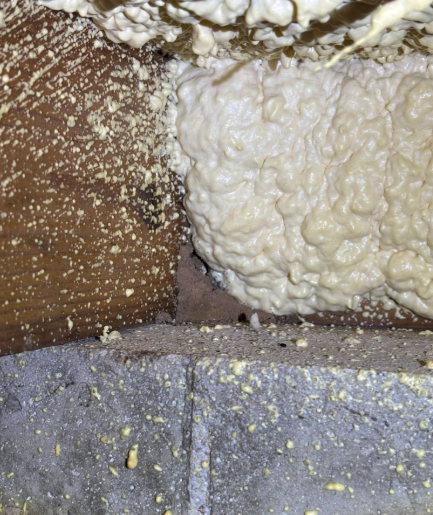Expanding spray foam insulation applied between a wooden support beam and a grey concrete foundation wall.