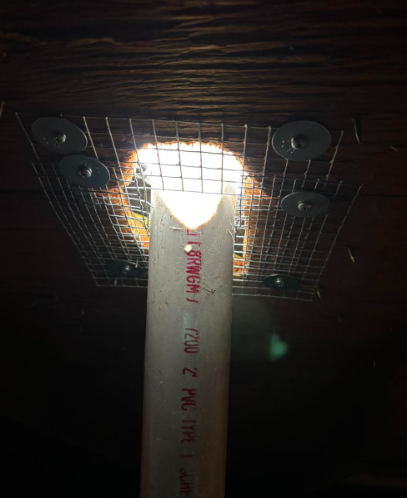 A vertical PVC pipe passes through a hole in a wooden ceiling, surrounded by a wire mesh guard held by washers.