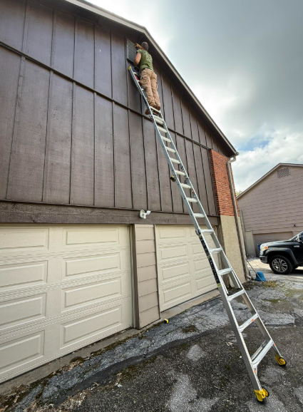A person stands on a long extension ladder leaning against the brown wood siding of a two-story residential garage.