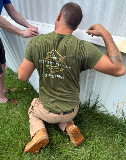 Two individuals in work attire kneel outdoors, collaboratively installing white siding on the side of a structure.