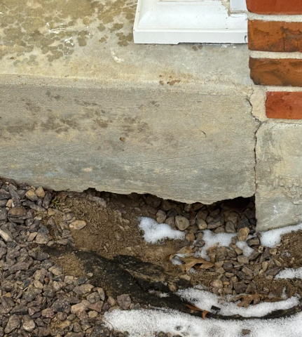 A concrete step has detached from the brick wall, leaving a hollow gap and cracked foundation above the gravel ground.