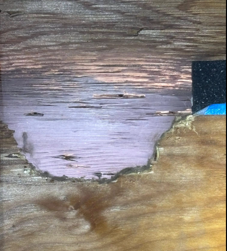 A patch of wood with a light purple, textured inlay surrounded by natural wood grain and a small black, blue-edged corner.
