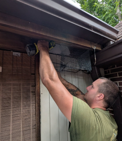 A person in a green t-shirt uses a power drill to install metal mesh near a roof eave on the exterior of a brick home.