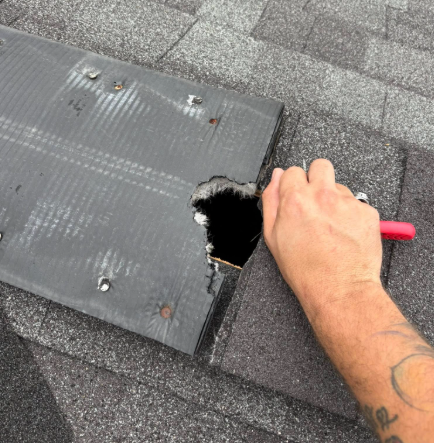 A hand holding a red tool inspects a hole in the roof shingles and underlying material.