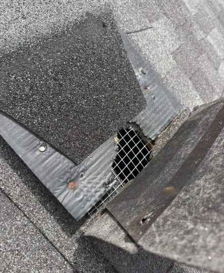 A damaged area of a shingled roof showing a hole covered by wire mesh, revealing dark space underneath.