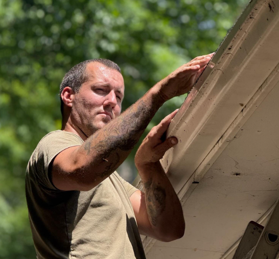 A person wearing a tan t-shirt reaches up to inspect or repair the edge of a light-colored roof outdoors.