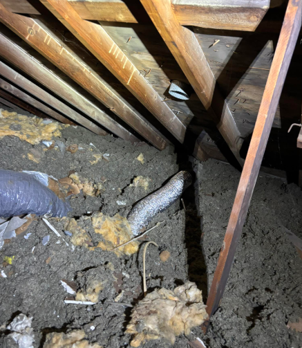 A dusty attic interior featuring wooden rafters, loose-fill insulation, and an exposed silver flexible air duct.