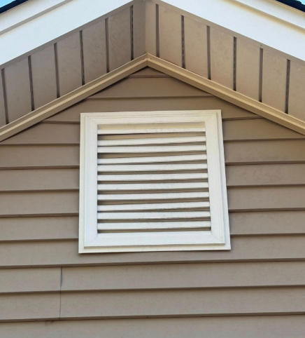 A square, white louvered gable vent centered on tan horizontal siding under a roof overhang.