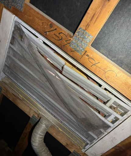 A damaged, dust-covered ventilation screen hanging from a wooden attic frame with a connected flexible duct hose.