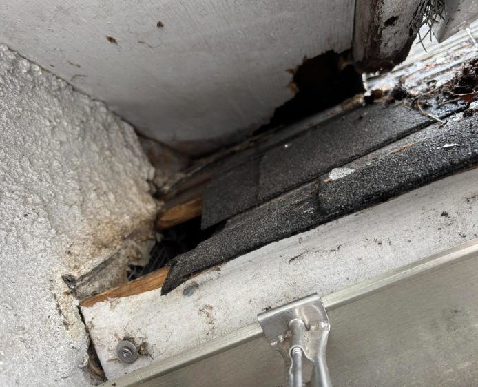 A close-up view of a damaged roof eave, showing rotting wood, missing shingles, and a gap against a textured wall.