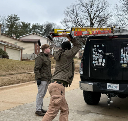 Two people working outside, one lifting a yellow and red ladder onto a black van rack while the other watches.