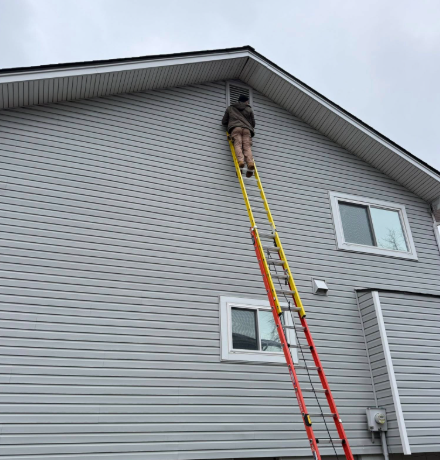 A person stands on a tall, multi-colored extension ladder leaned against the side of a grey, vinyl-sided house.