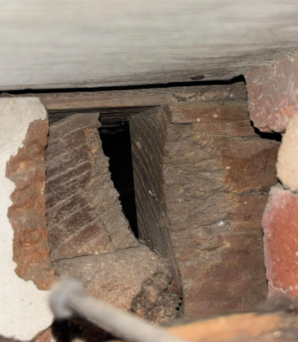 A close-up view of a gap in a brick wall revealing dark, weathered wooden structural beams behind the masonry.