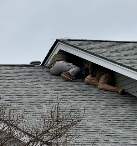 Two people crawl into a dark attic space through an open gable vent on a shingled roof.