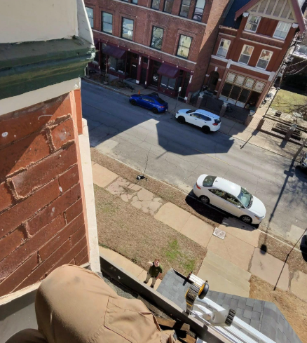 An elevated view looking down from a brick building at a person standing on a sidewalk near parked cars on a city street.