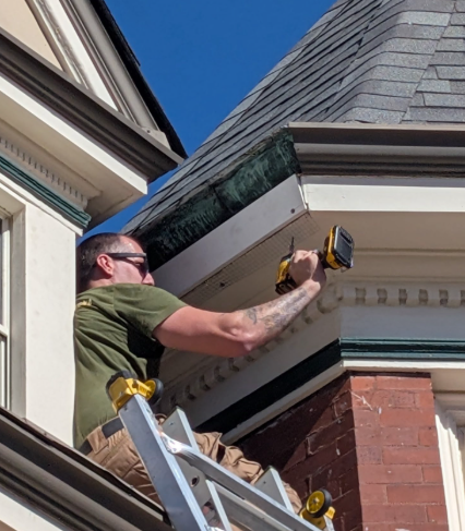 A person on a ladder uses a power drill to work on the exterior trim of a building with a shingled turret roof.