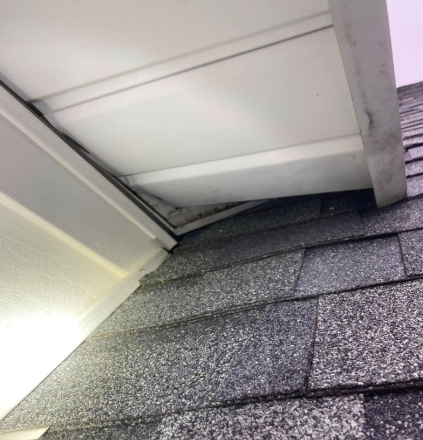 White vinyl soffit panels hanging detached from the eaves of a roof above dark gray asphalt shingles.