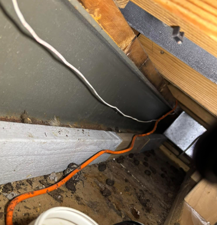 An orange extension cord and white wire run across a dusty attic floor near wooden beams and metallic air ducts.