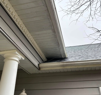 A low-angle view of a house exterior showing a white pillar, soffit, and roof shingles meeting the gutter.