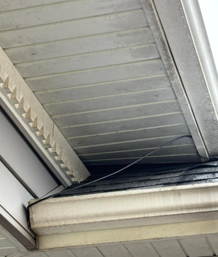A loose black cable hangs from the white soffit of a home, dangling over the dark shingles of the roofline.