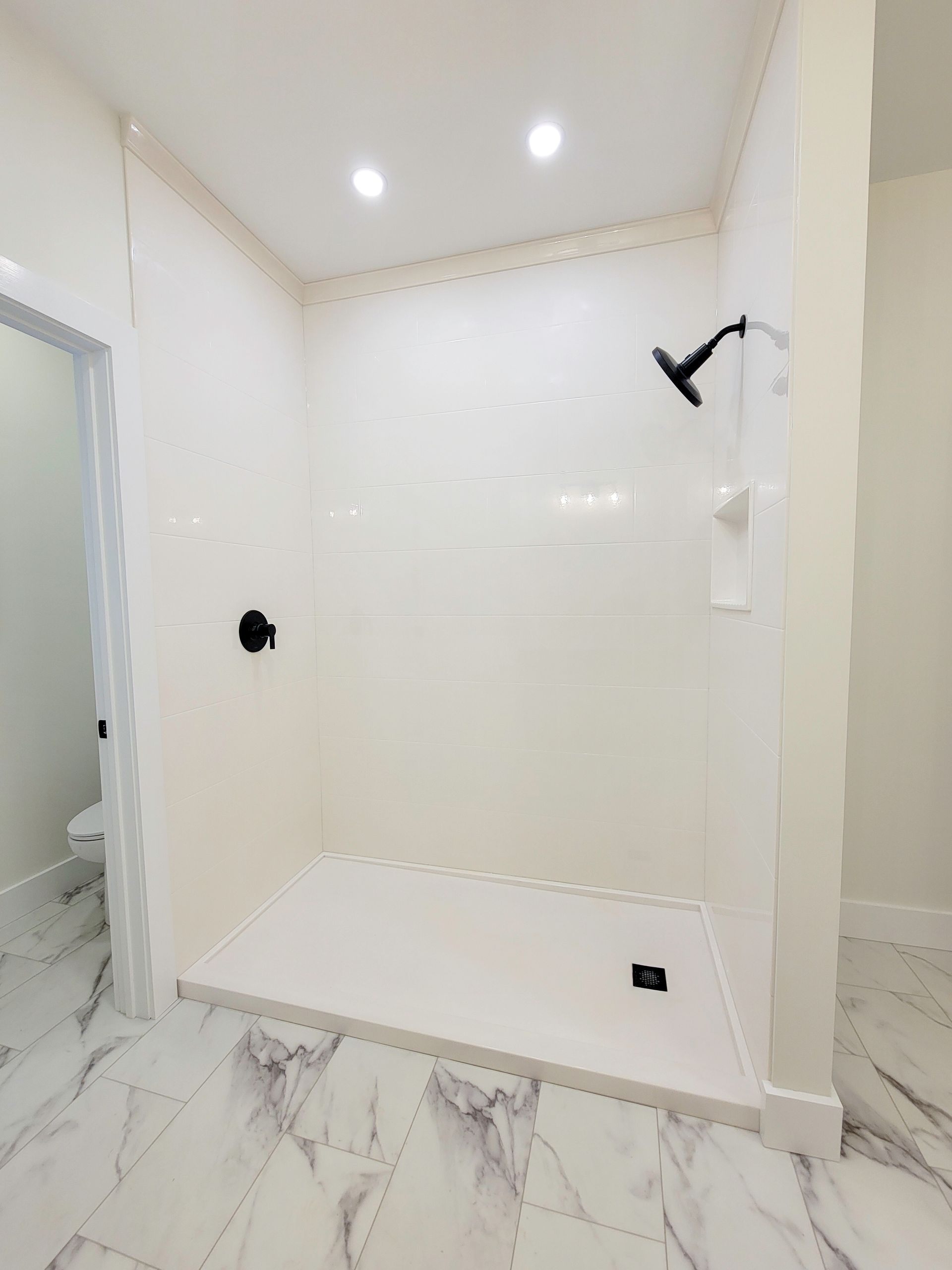 Empty modern shower stall with white tile, marble-look floor, wall niche, and black shower fixtures