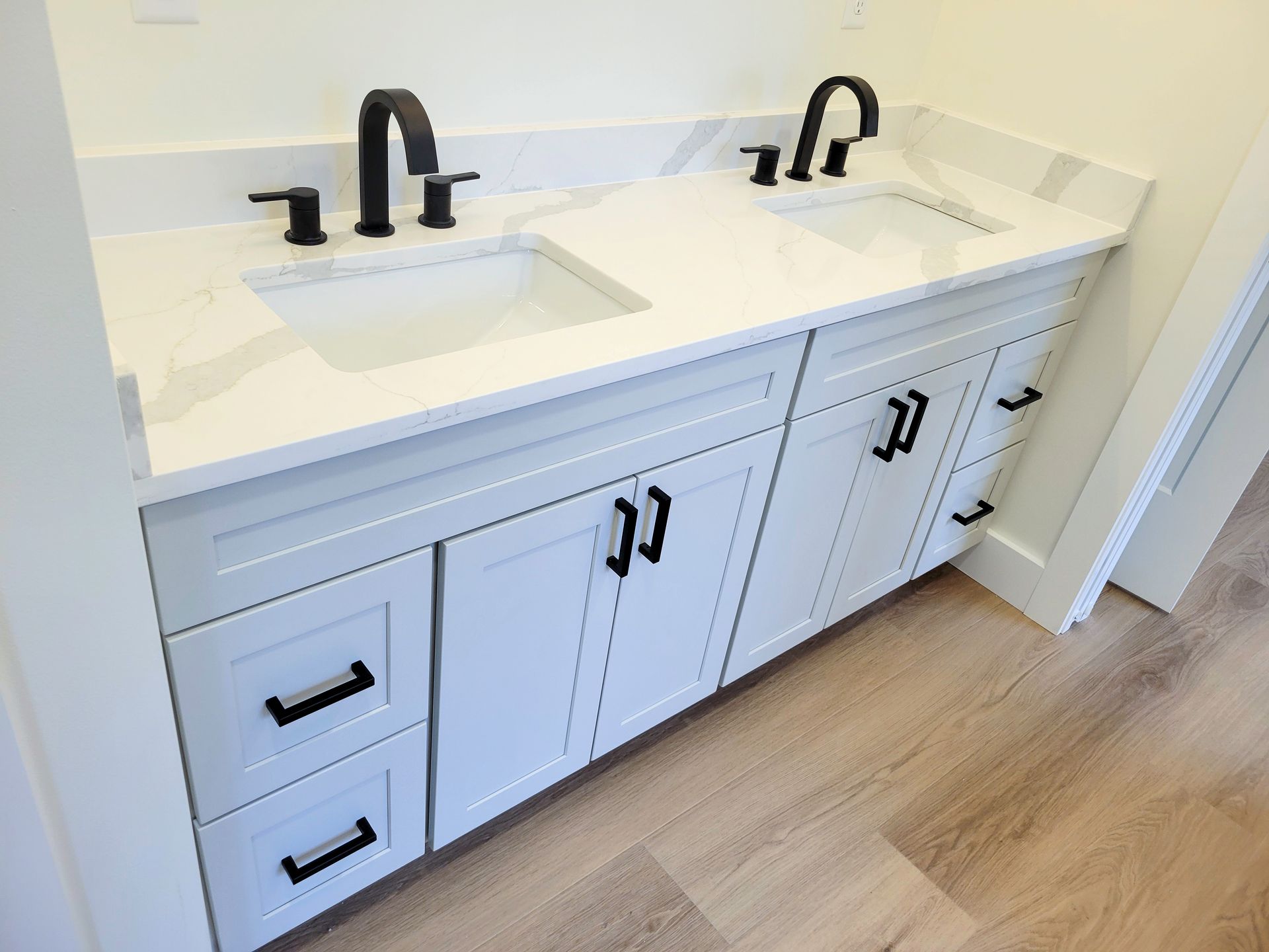 Double vanity with white countertop, two sinks, black faucets, and light wood flooring