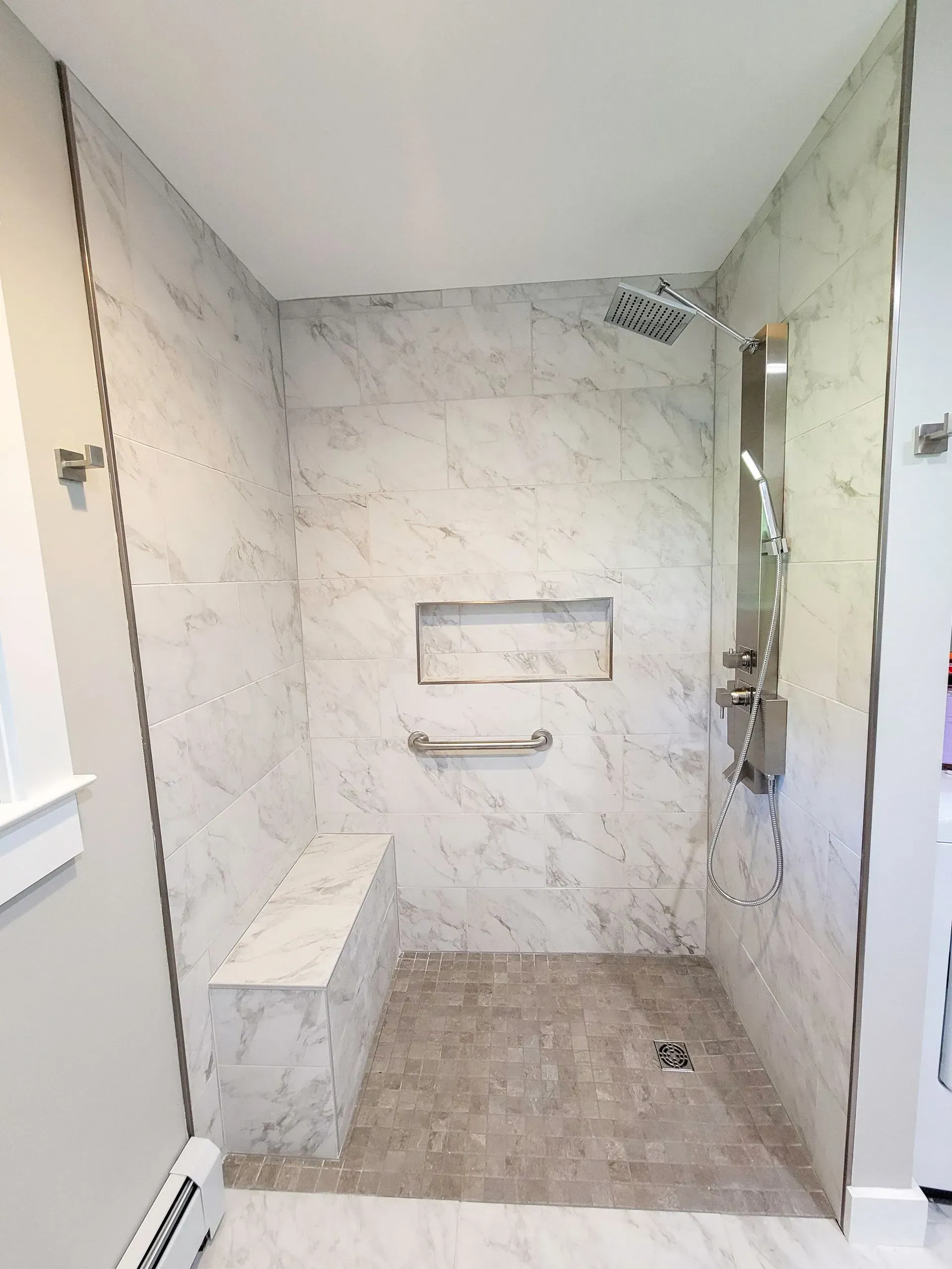 Newly constructed accessible shower with marble-look tile, built-in seat, and grab bar.