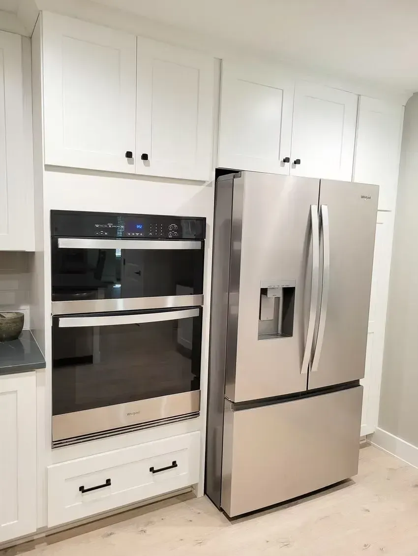Stainless steel fridge next to a built-in oven and white cabinets in a kitchen.