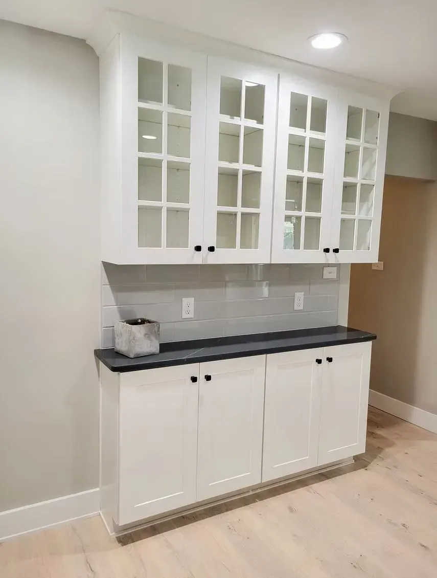 White cabinets with glass-front uppers and black countertop. A gray backsplash and black cabinet knobs are also present.
