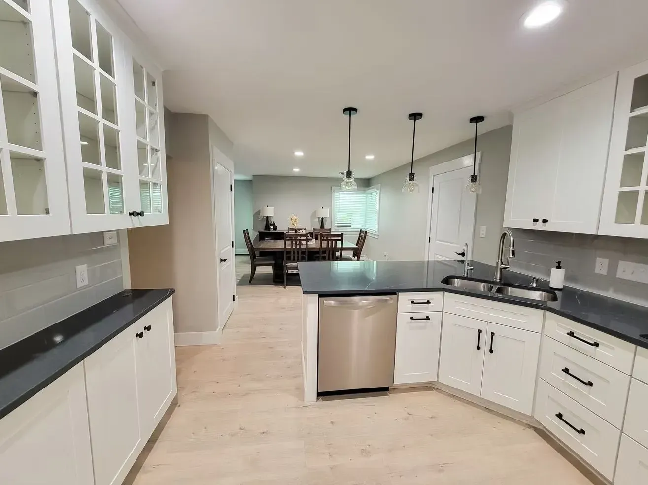 Modern kitchen with white cabinets, dark countertops, and stainless steel appliances. Dining room visible in the background.