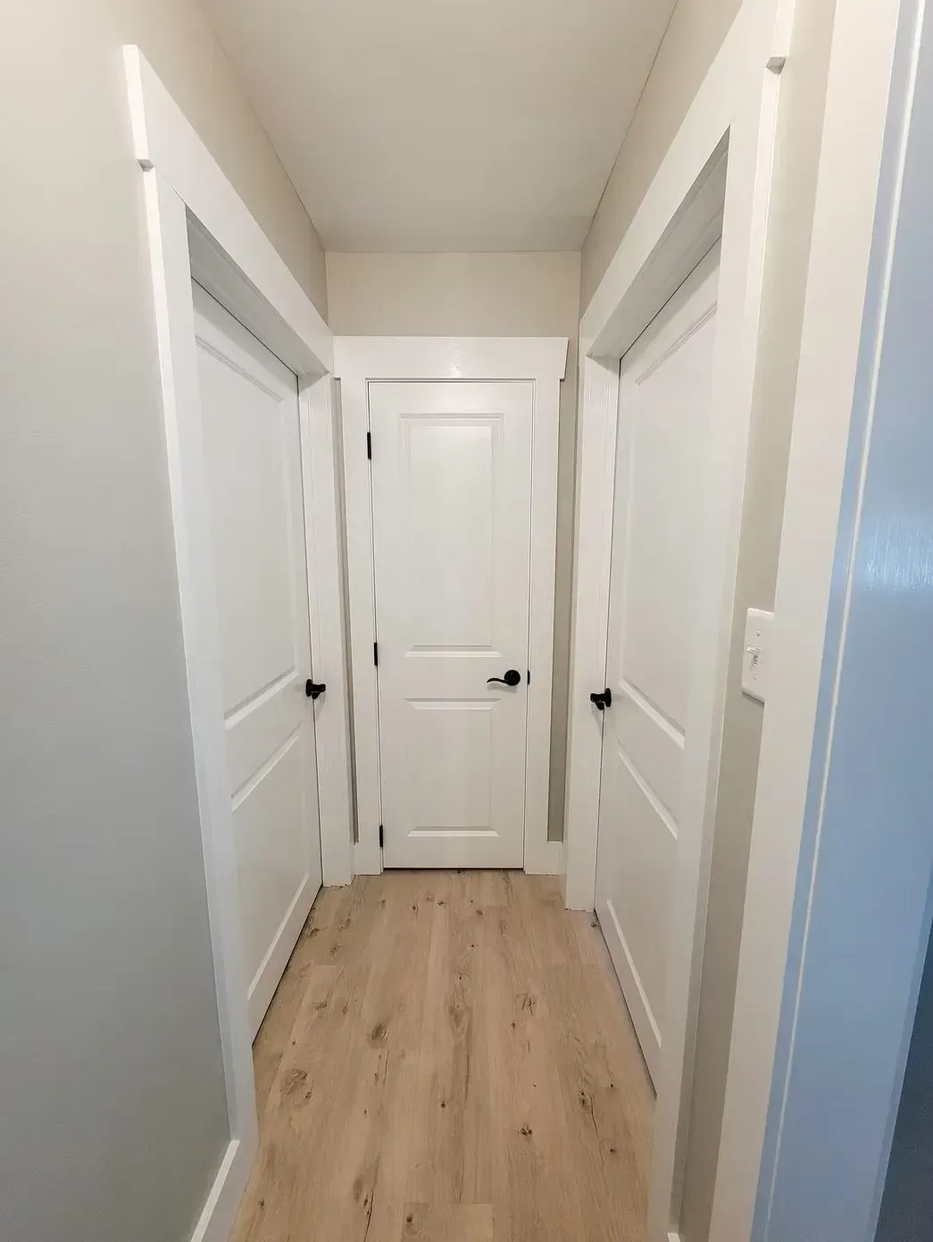 Narrow hallway with three white doors, light wood-look floor, and gray walls.