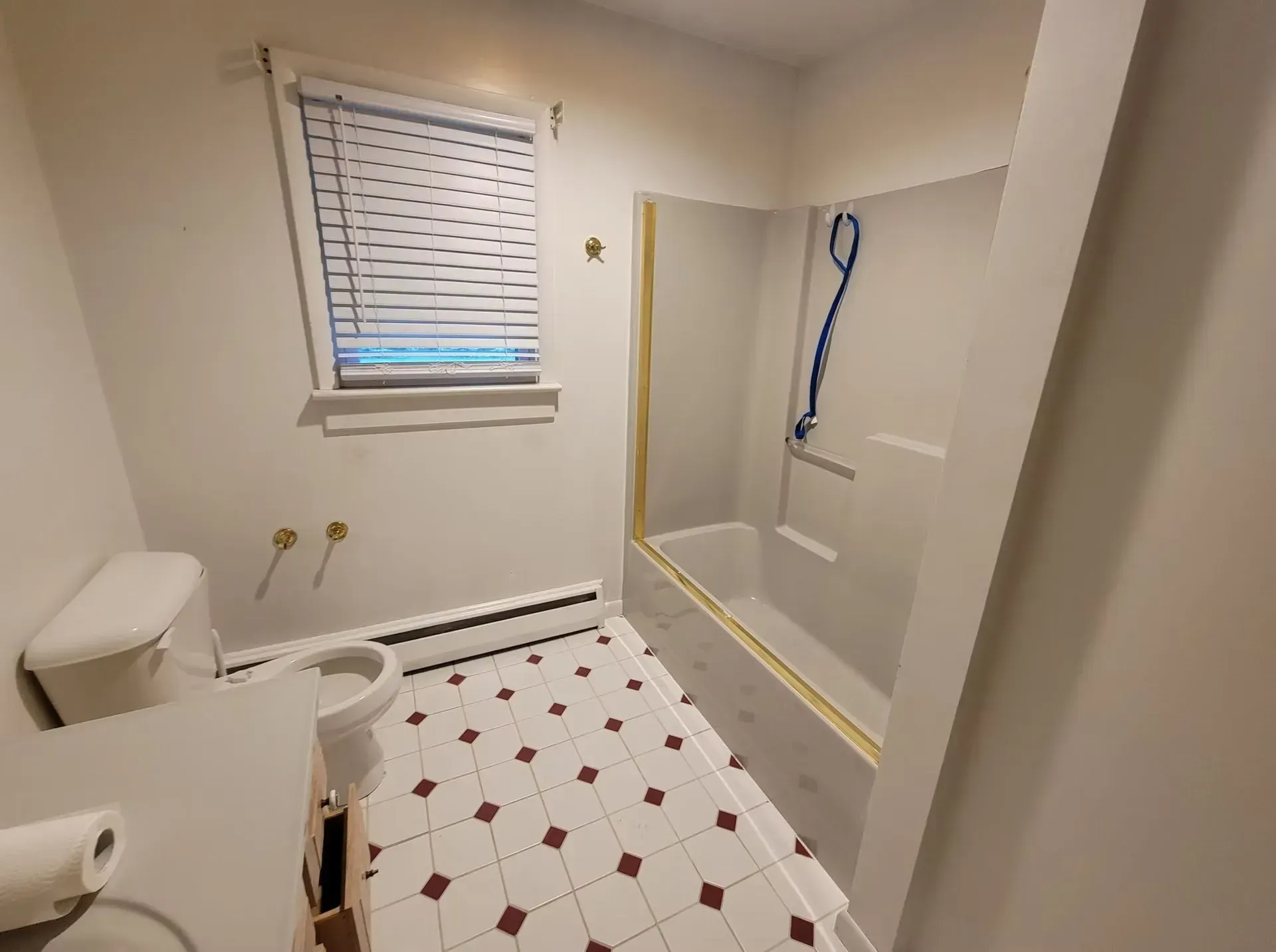 Bathroom with toilet, vanity, and shower/tub. White walls, red and white checkered floor, window with blinds.