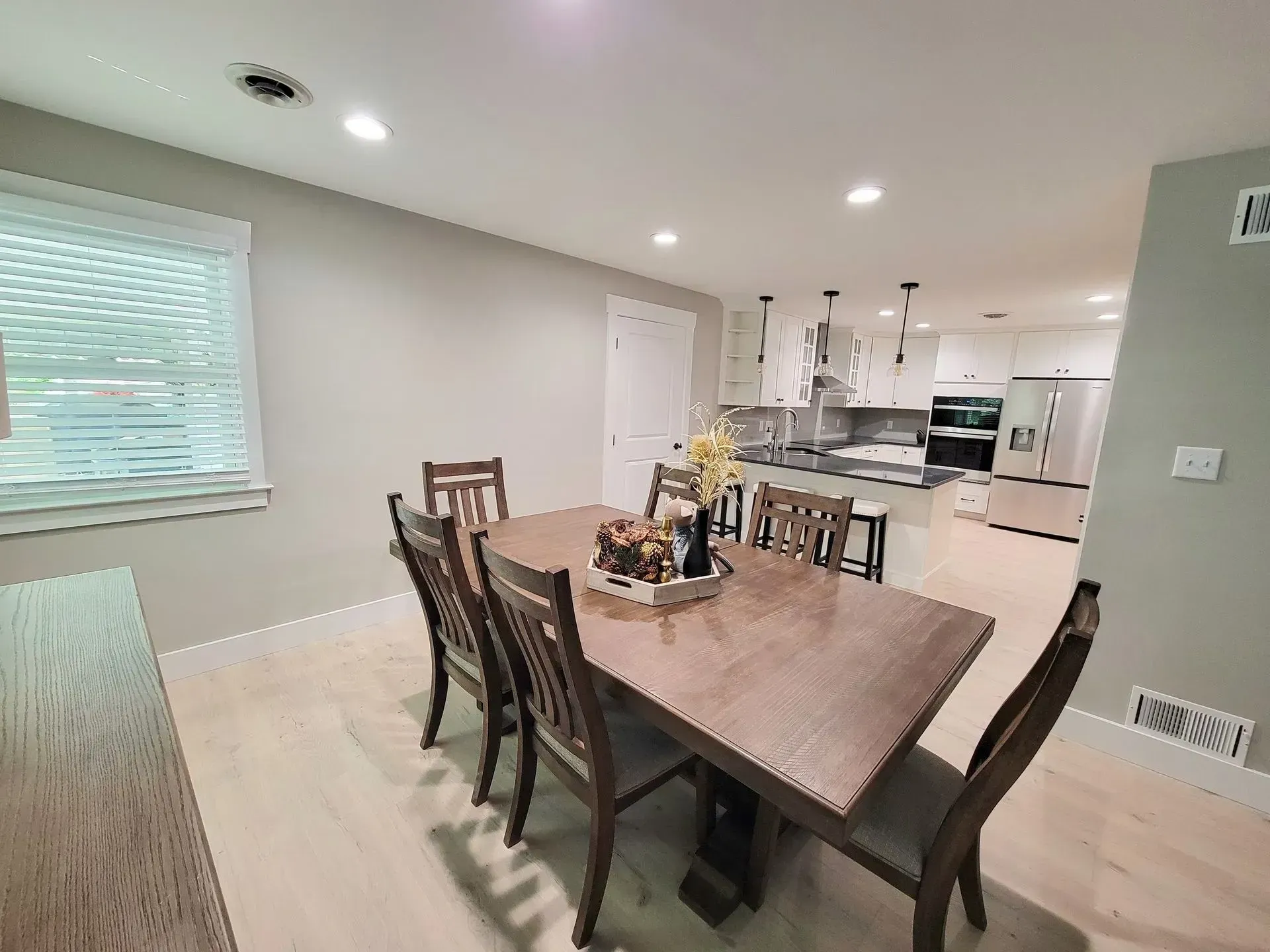 Dining room with wooden table and chairs, light flooring, and open kitchen in the background.