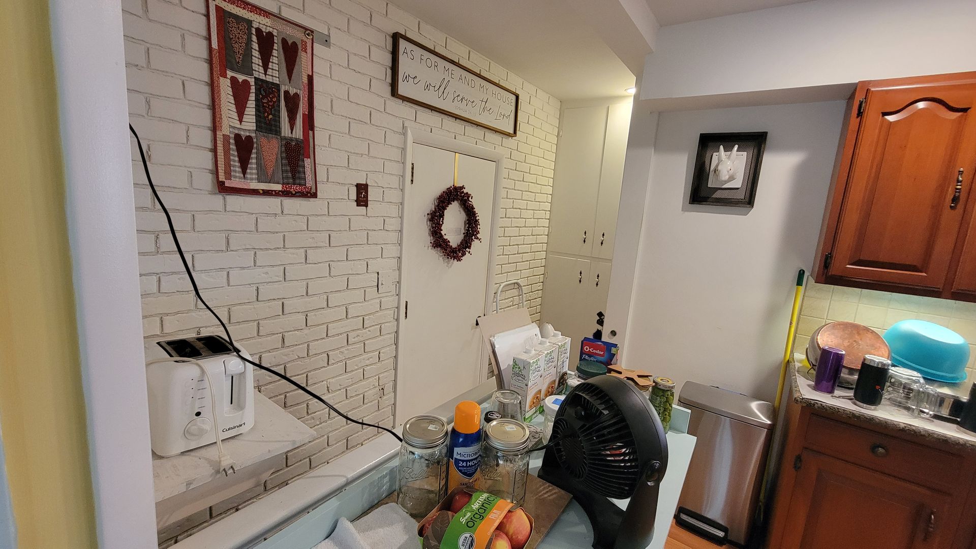 Kitchen with white brick wall, door, and wooden cabinets.  Counter with toaster, food, and stainless steel trash can.