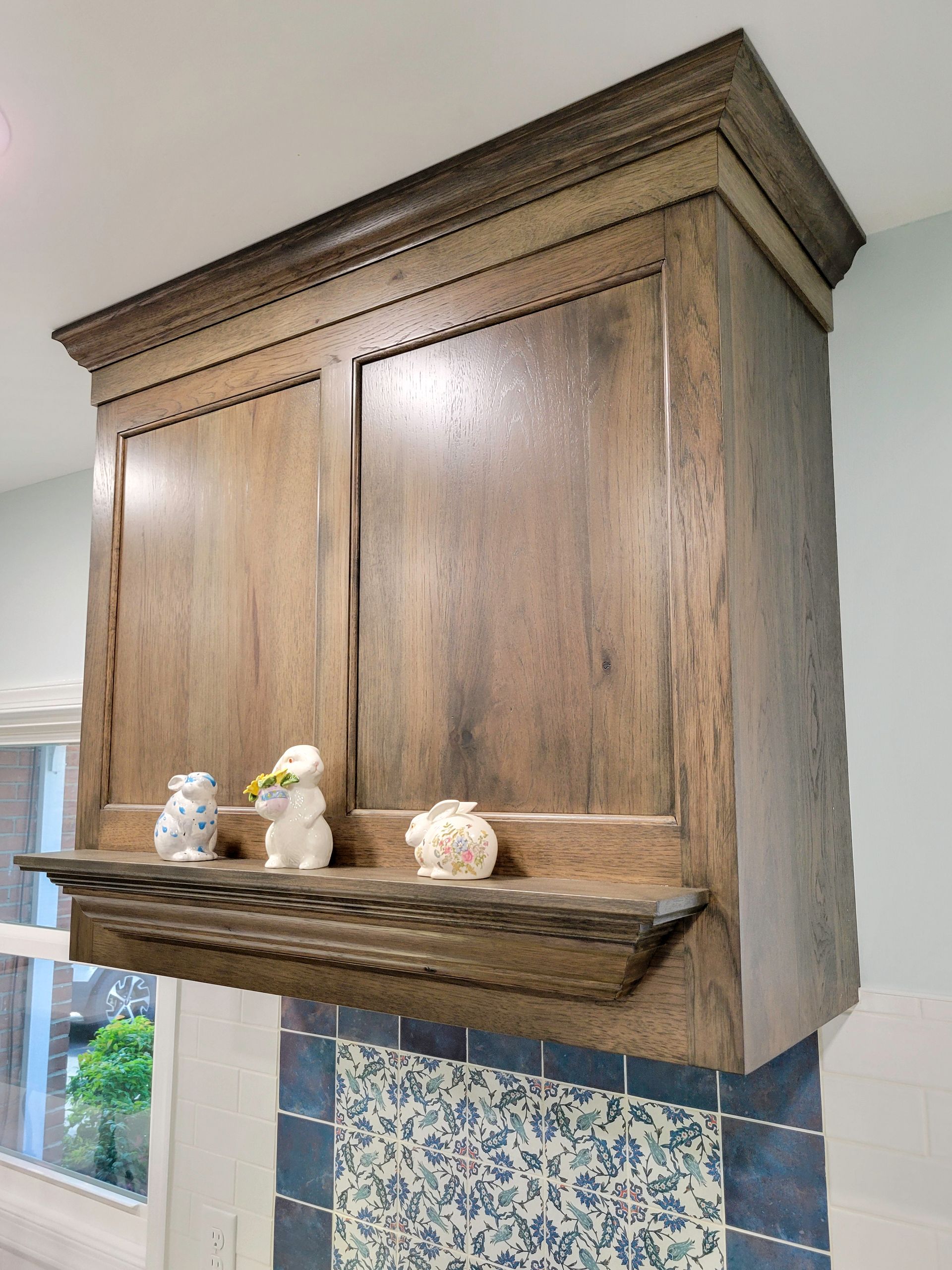 Wooden kitchen cabinet with shelf holding figurines above blue tile backsplash.